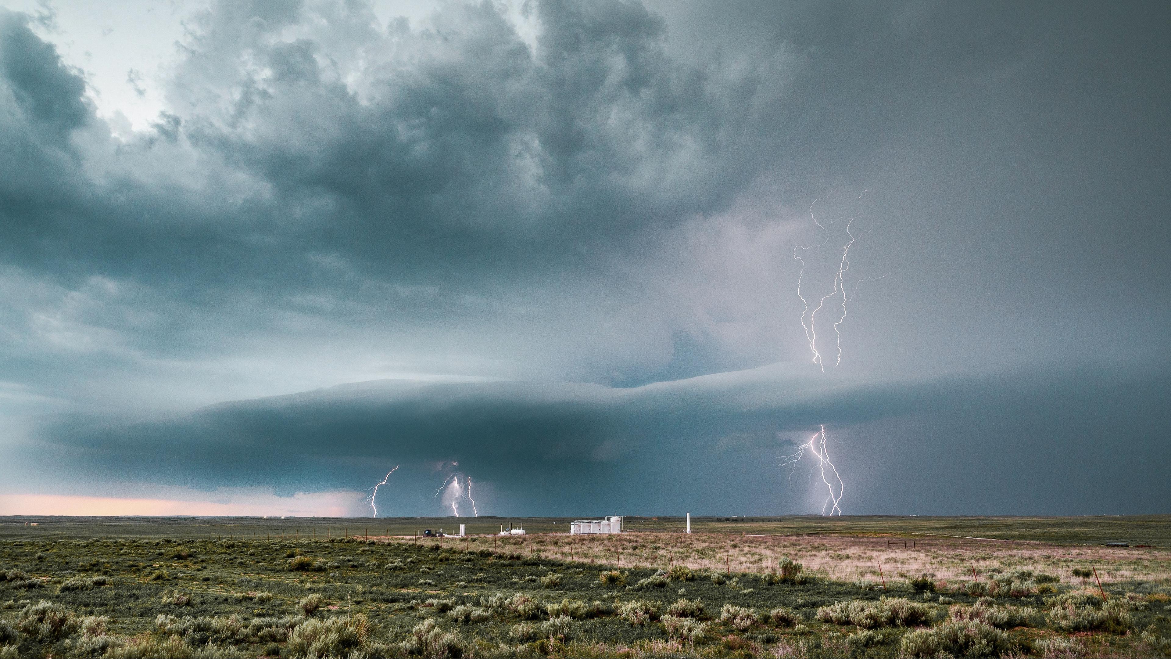 A vast landscape under a dark, stormy sky with multiple lightning bolts striking above open fields and distant structures.