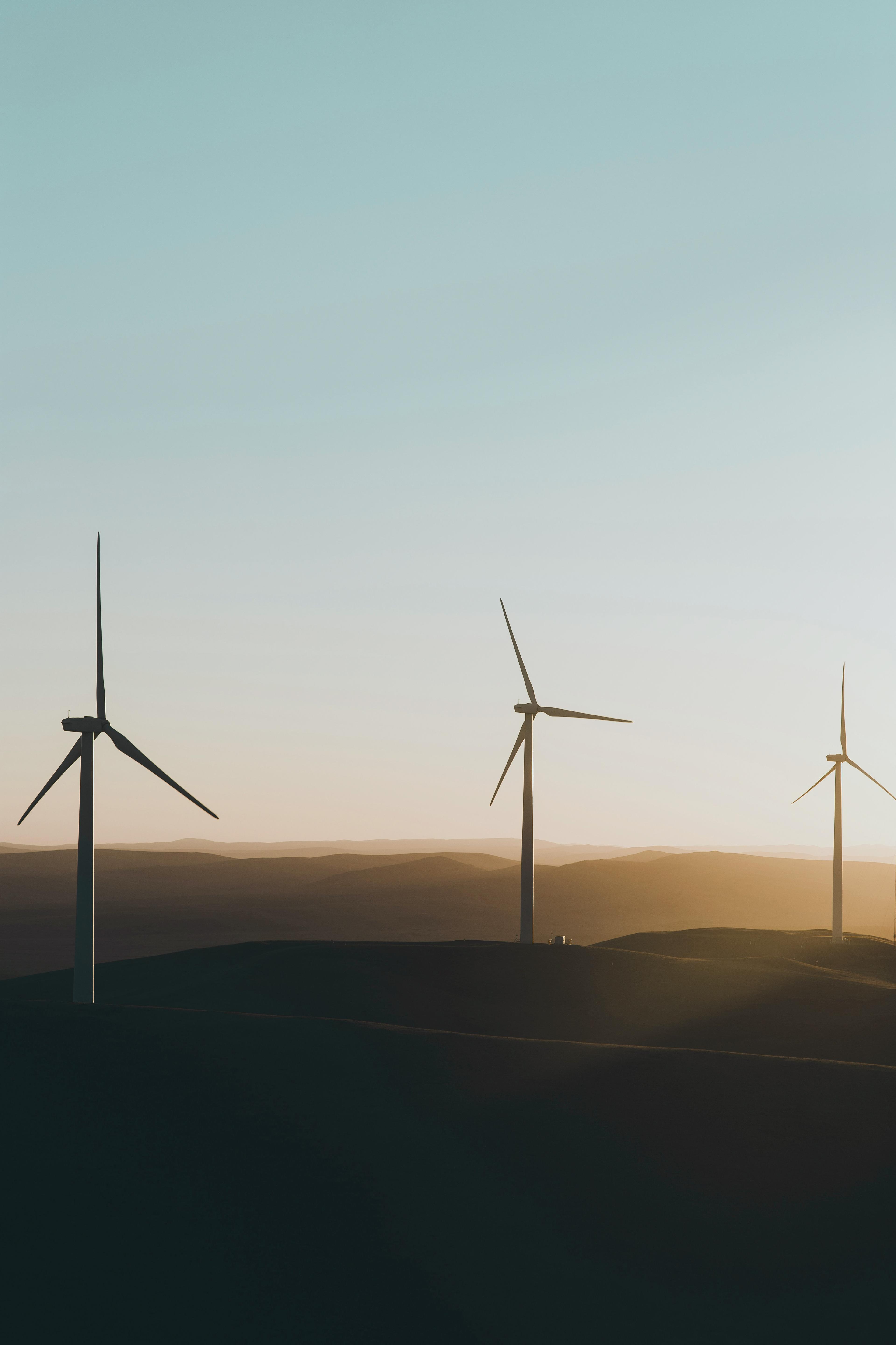Three wind turbines stand on rolling hills under a clear sky at sunrise, casting long shadows across the landscape.
