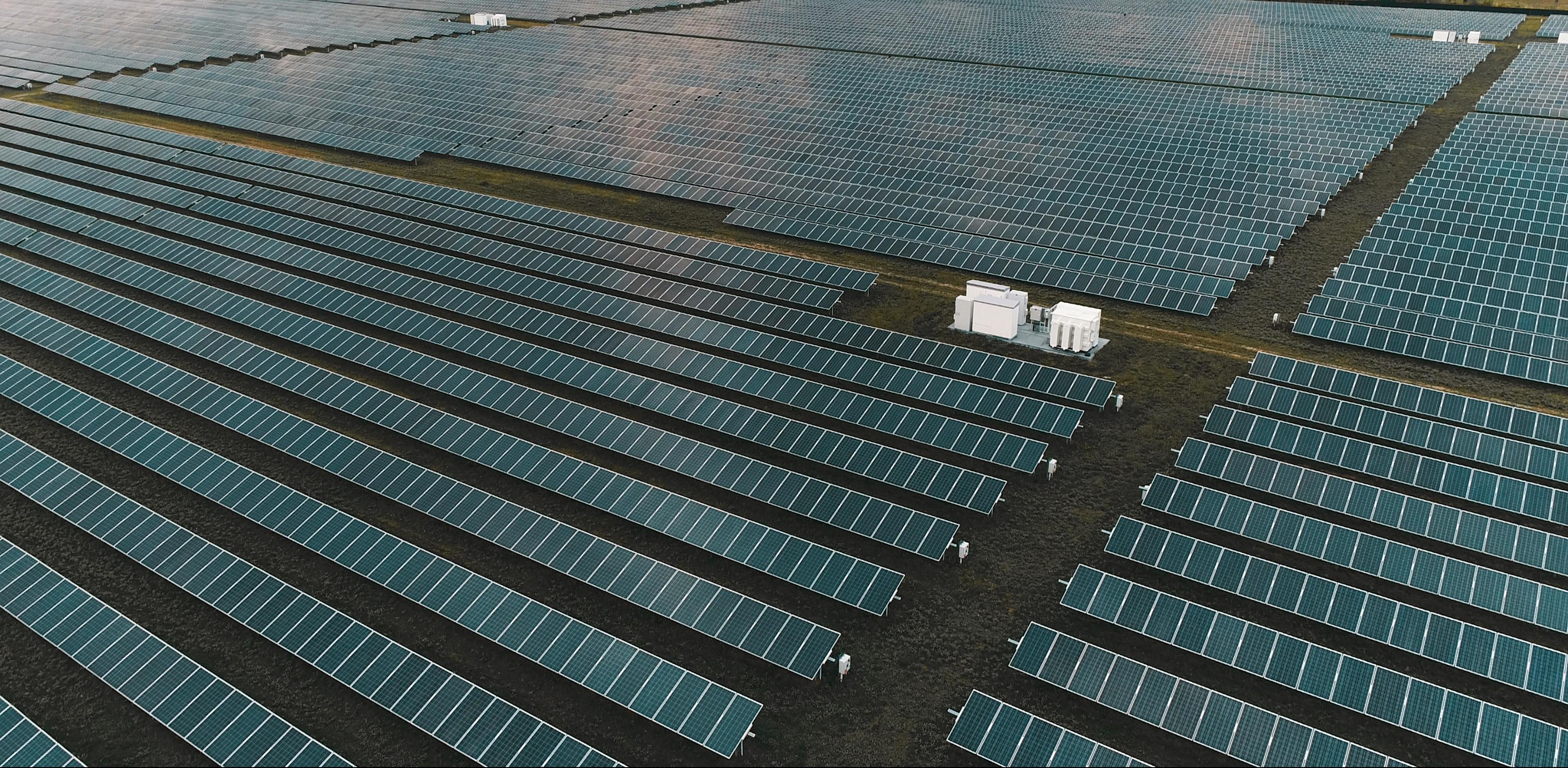 Aerial view of a vast solar farm with rows of solar panels on grassy terrain, accompanied by a cluster of white utility structures.