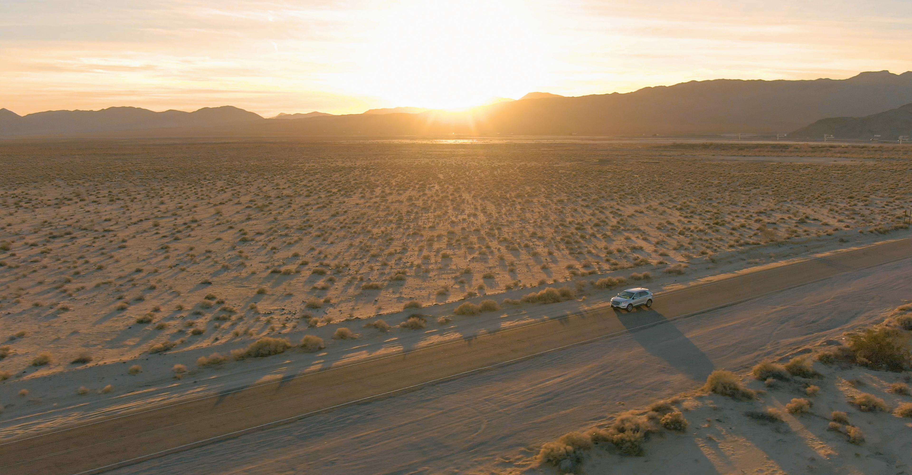 A car drives on a desert road at sunset, surrounded by sparse vegetation and distant mountains under a clear sky.