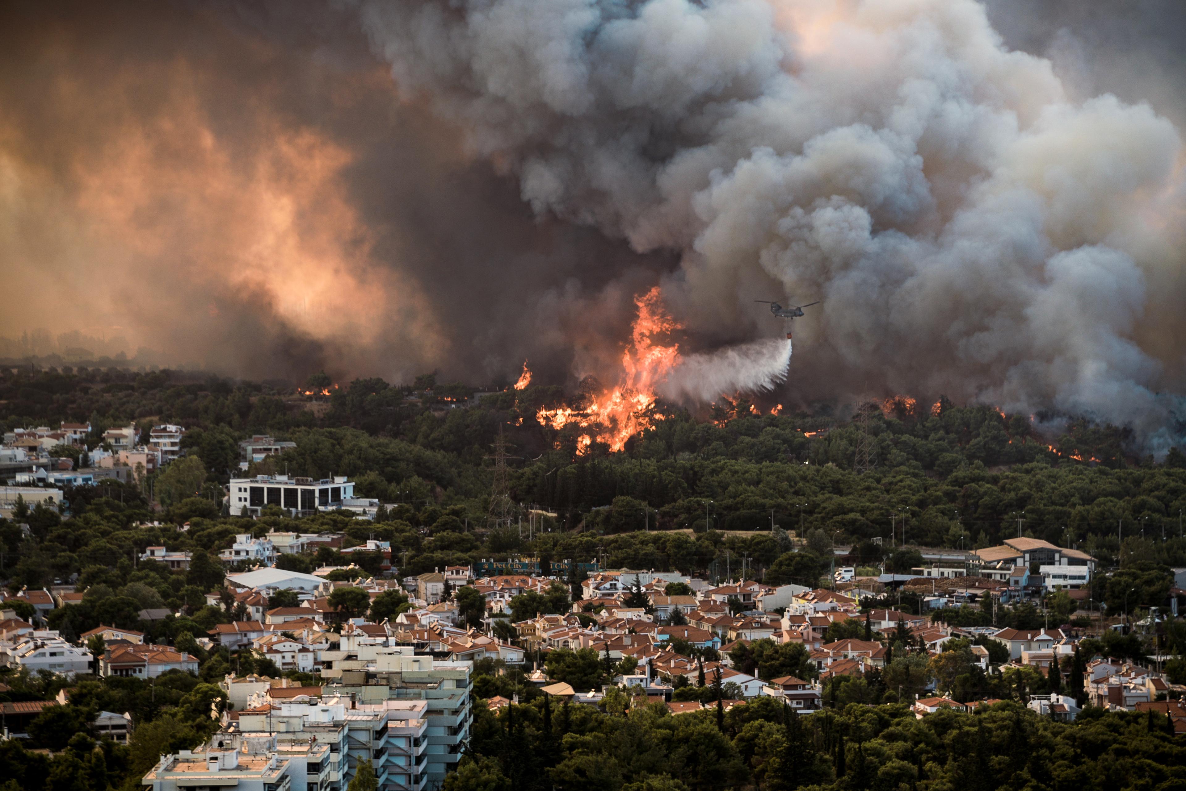 Aerial view of a wildfire near a residential area, with thick smoke and flames in the forest, and a firefighting plane overhead.
