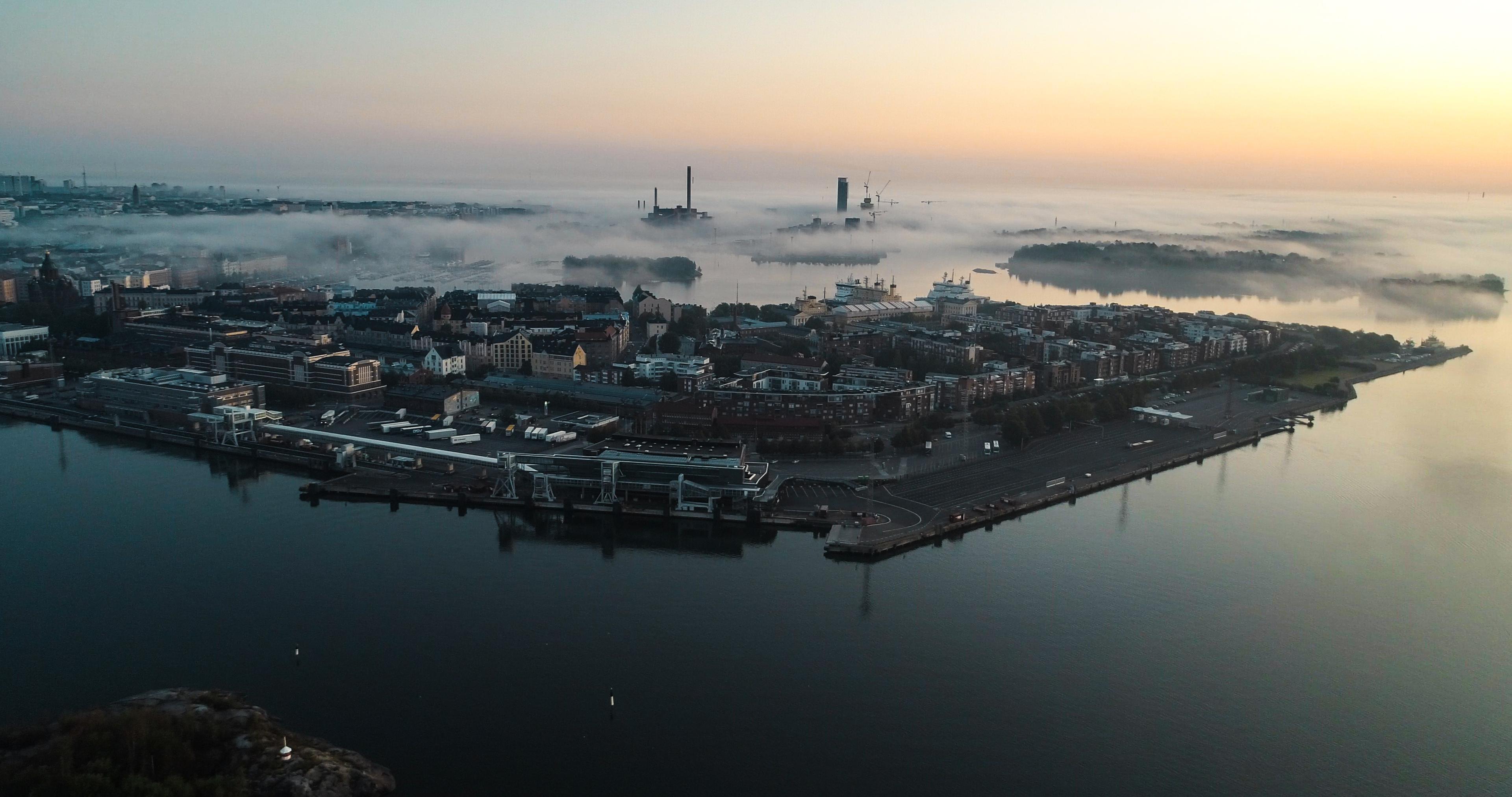 Aerial view of a coastal city at sunrise, with a port, industrial structures, fog over the water, and distant islands.