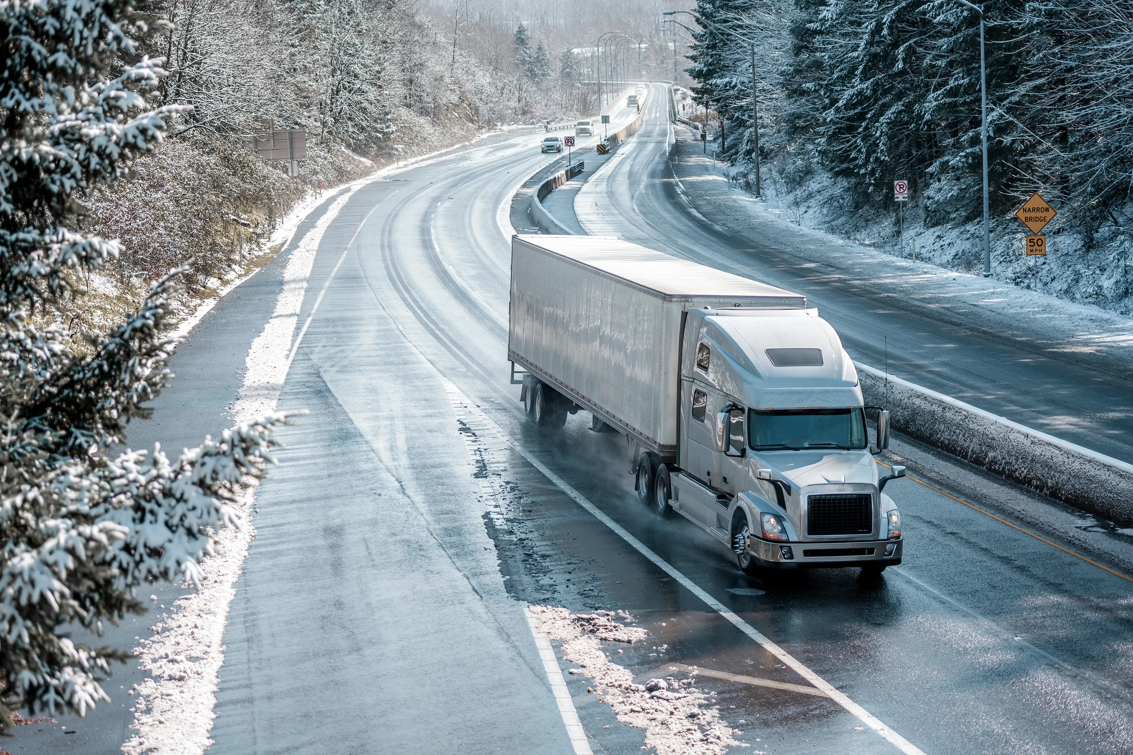A semi-truck drives on a snow-covered road through a wintry forest landscape, with cars in the distance and pine trees lining the road.