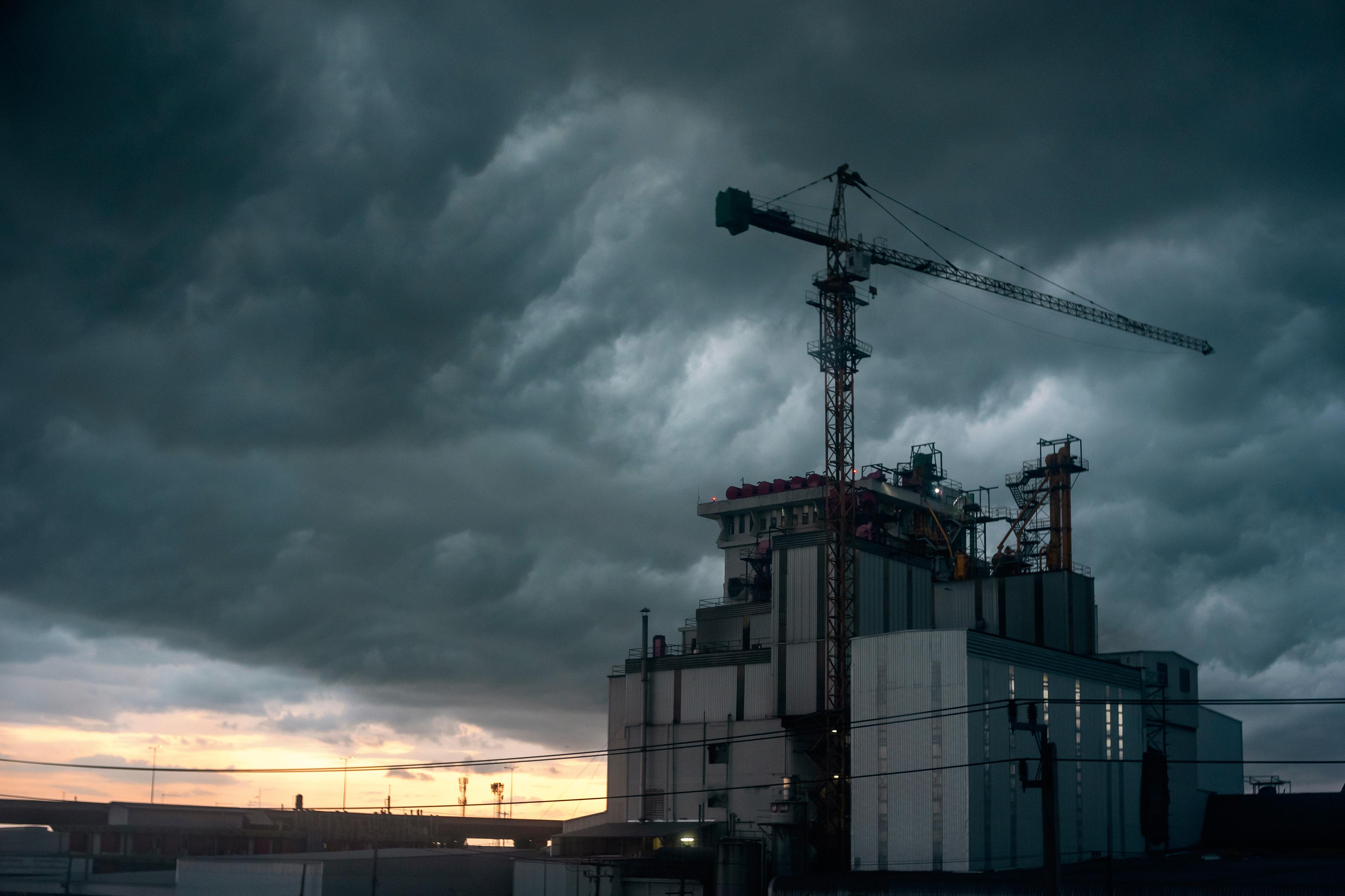 Industrial building with a crane under a dramatic, cloudy sky at dusk, with a hint of sunset on the horizon.