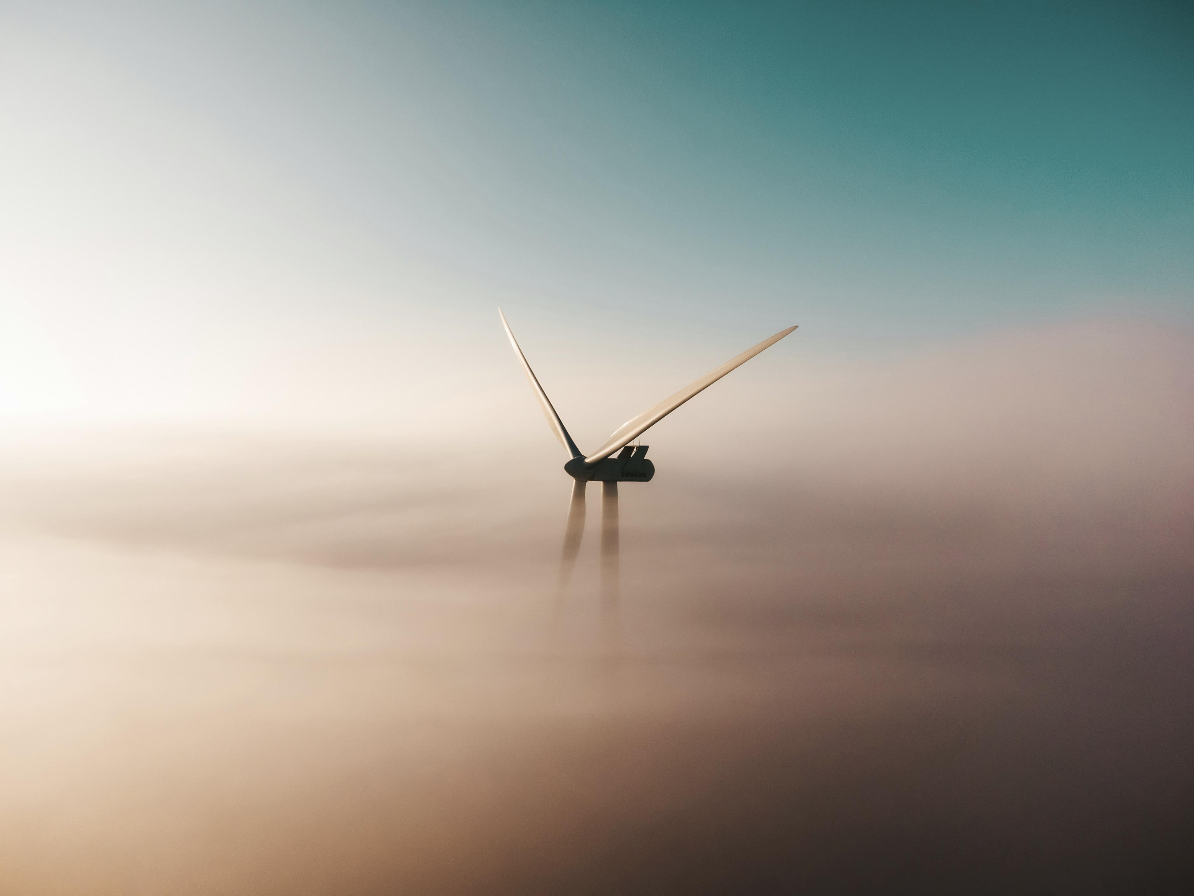 A solitary wind turbine emerges through thick fog under a clear blue sky.