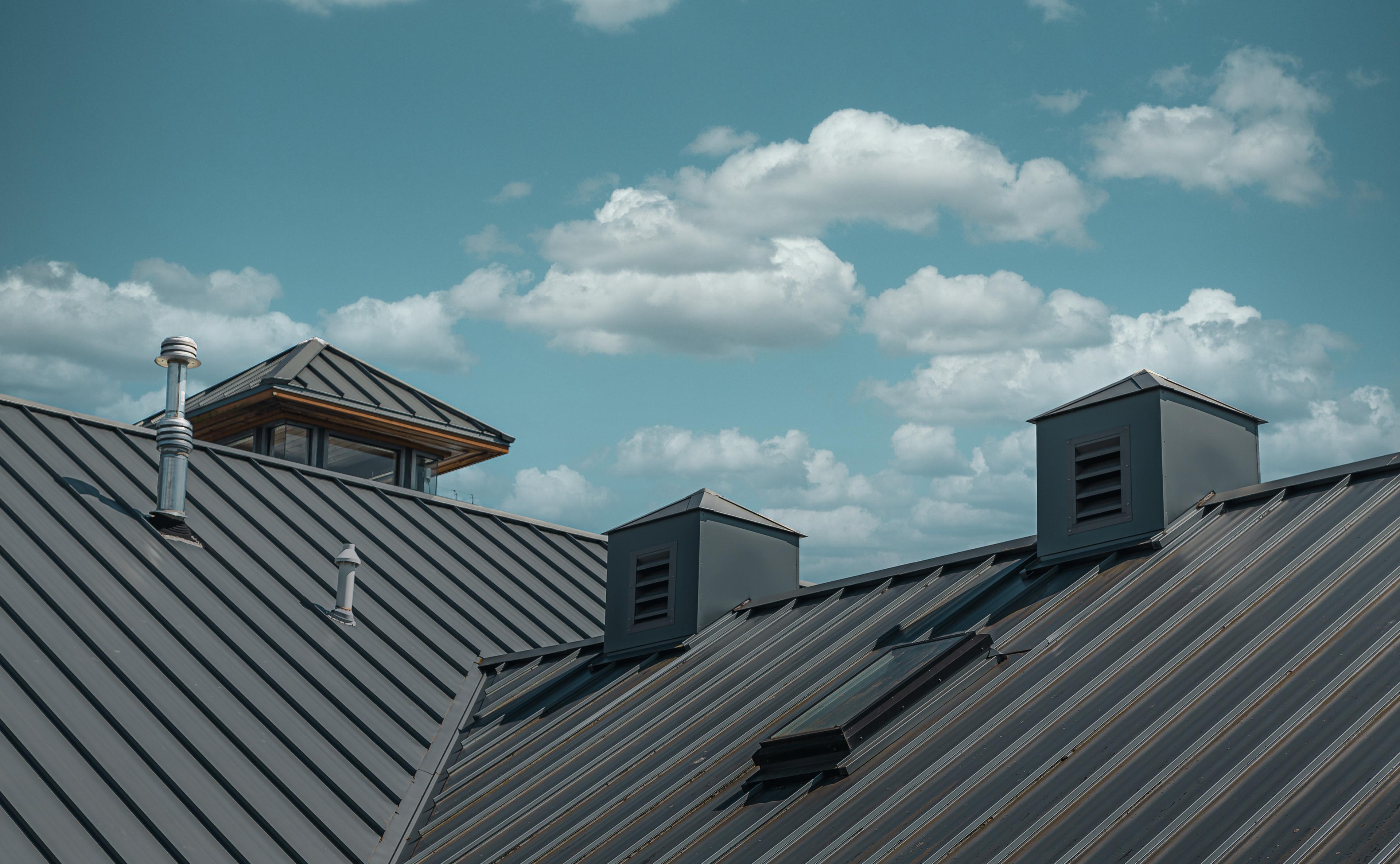 Metal roof with vents and chimneys under a blue sky with scattered clouds.