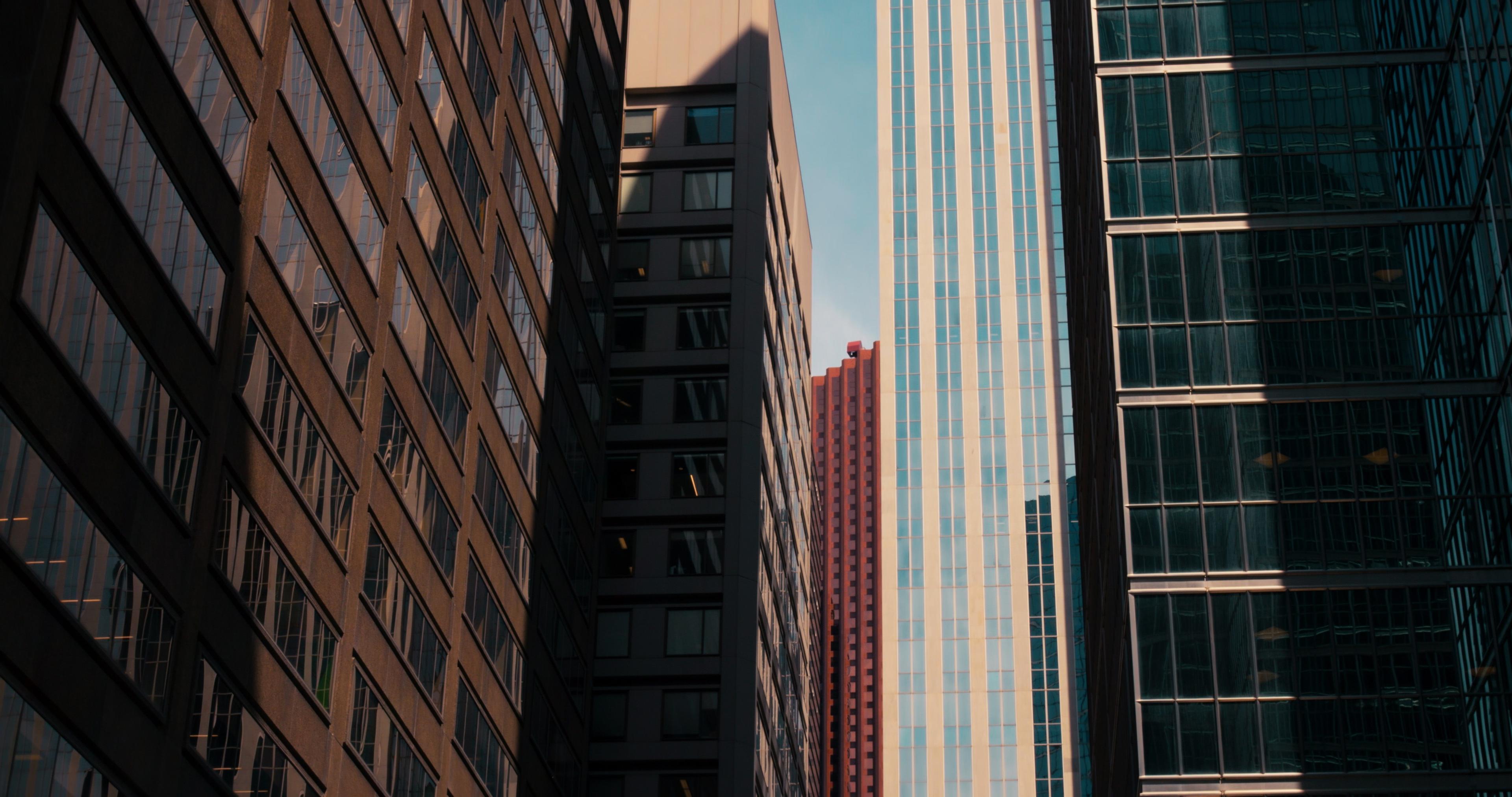 Tall, modern buildings with glass and steel facades rise into a clear blue sky, casting shadows on each other.