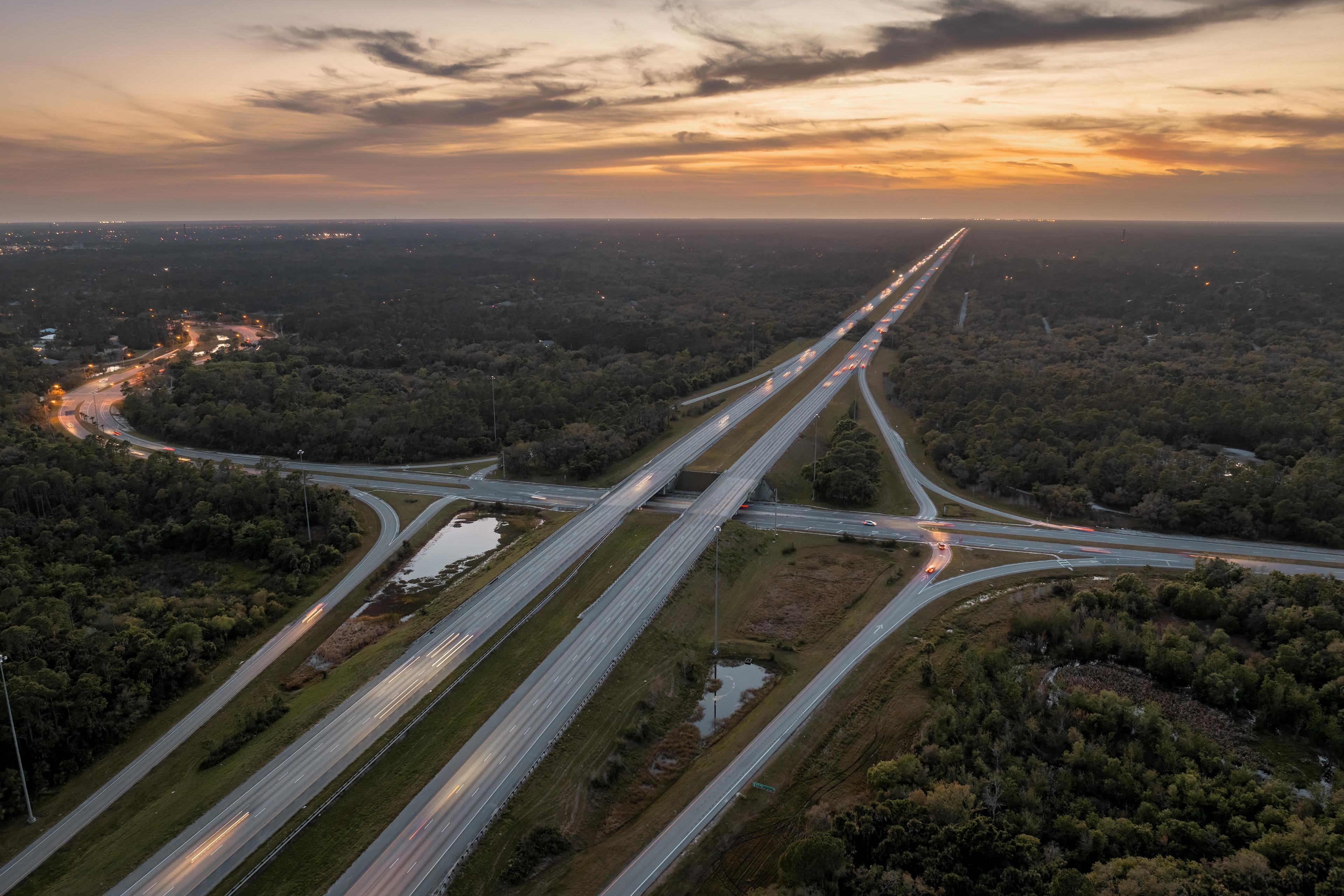 Aerial view of a highway intersection at sunset with light trails from cars, surrounded by lush greenery and a glowing sky.