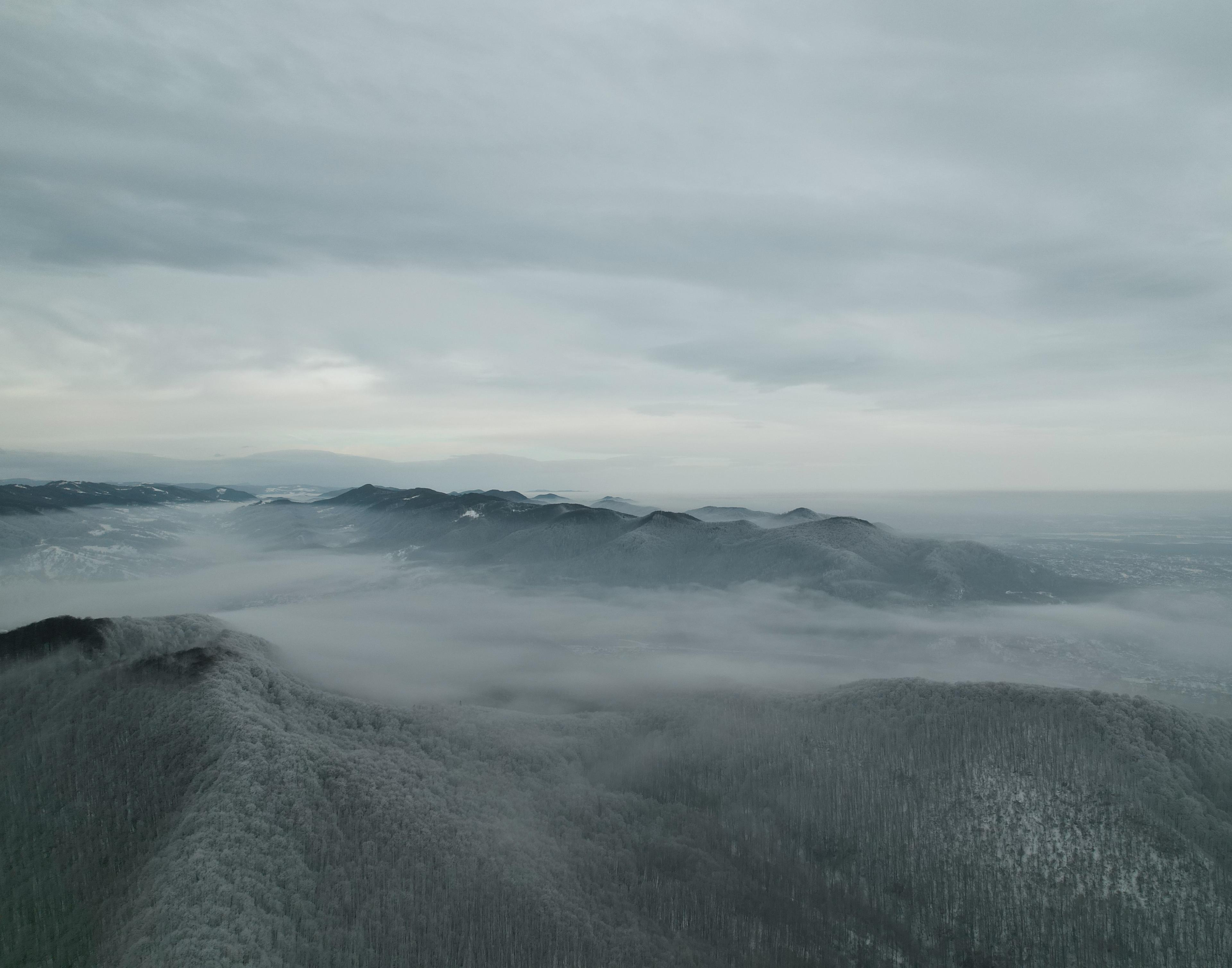 Foggy mountain landscape with snow-dusted ridges under an overcast sky, blending into distant valleys and hills.