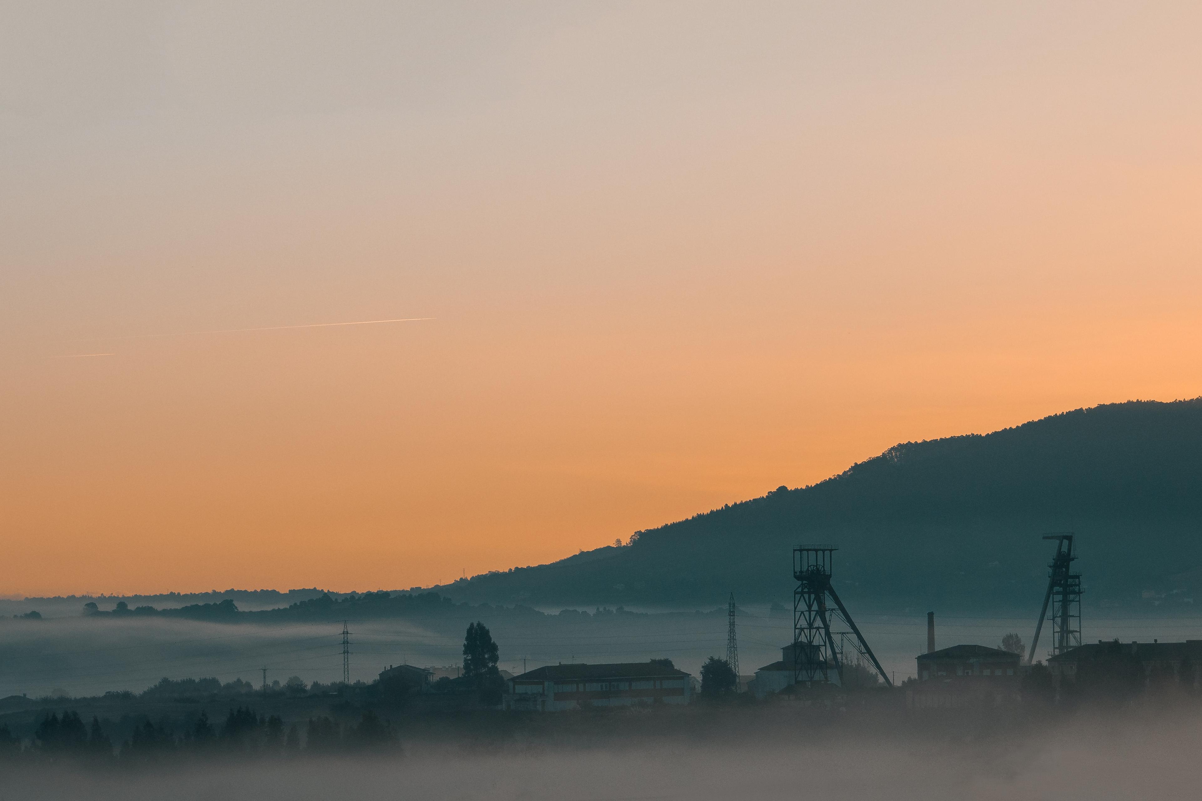 A foggy landscape at sunrise with distant industrial structures and a mountain silhouette under an orange sky.