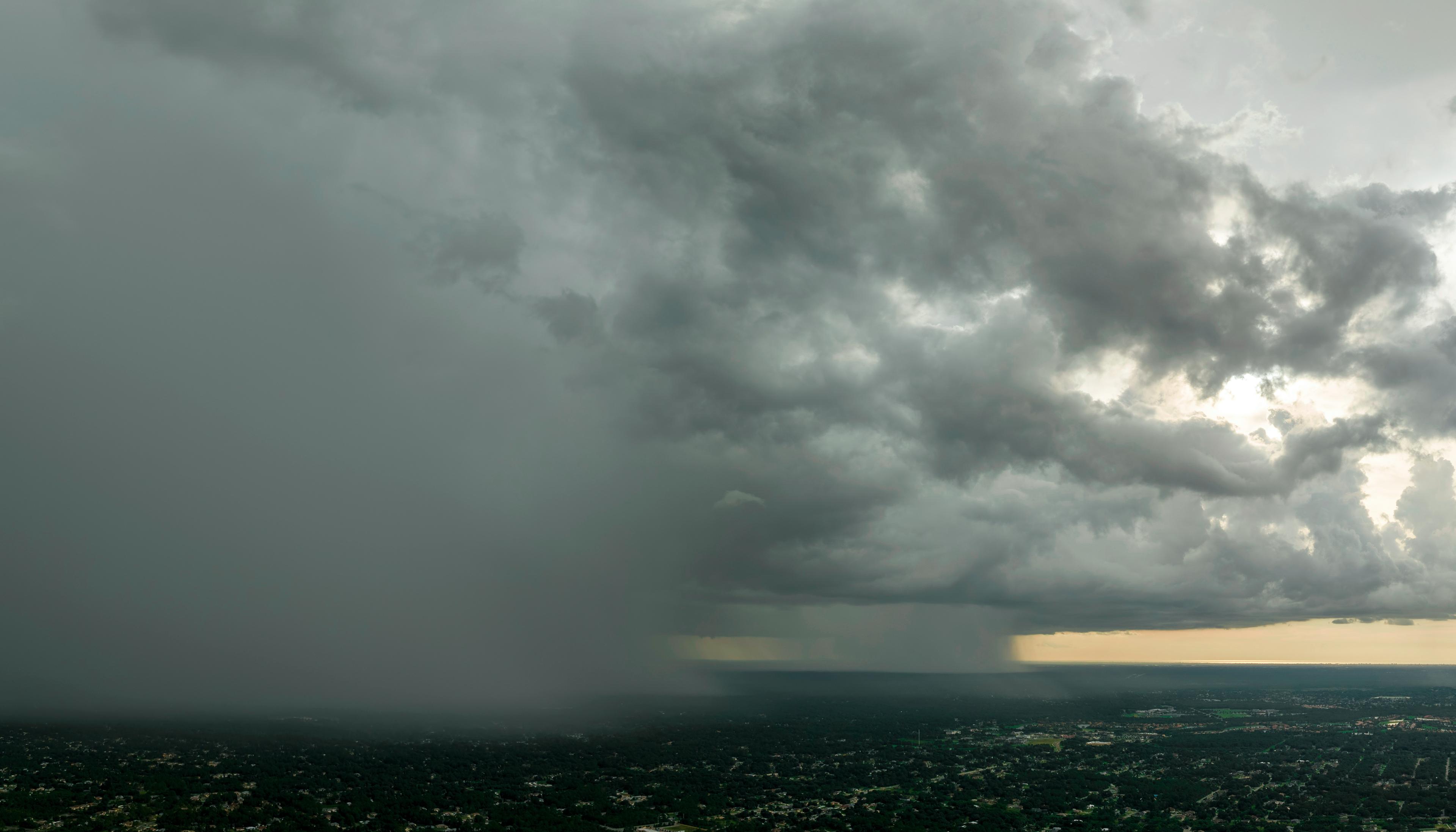 Aerial view of a city under a dramatic, dark storm cloud, with rain visible on the left and a lighter horizon on the right.