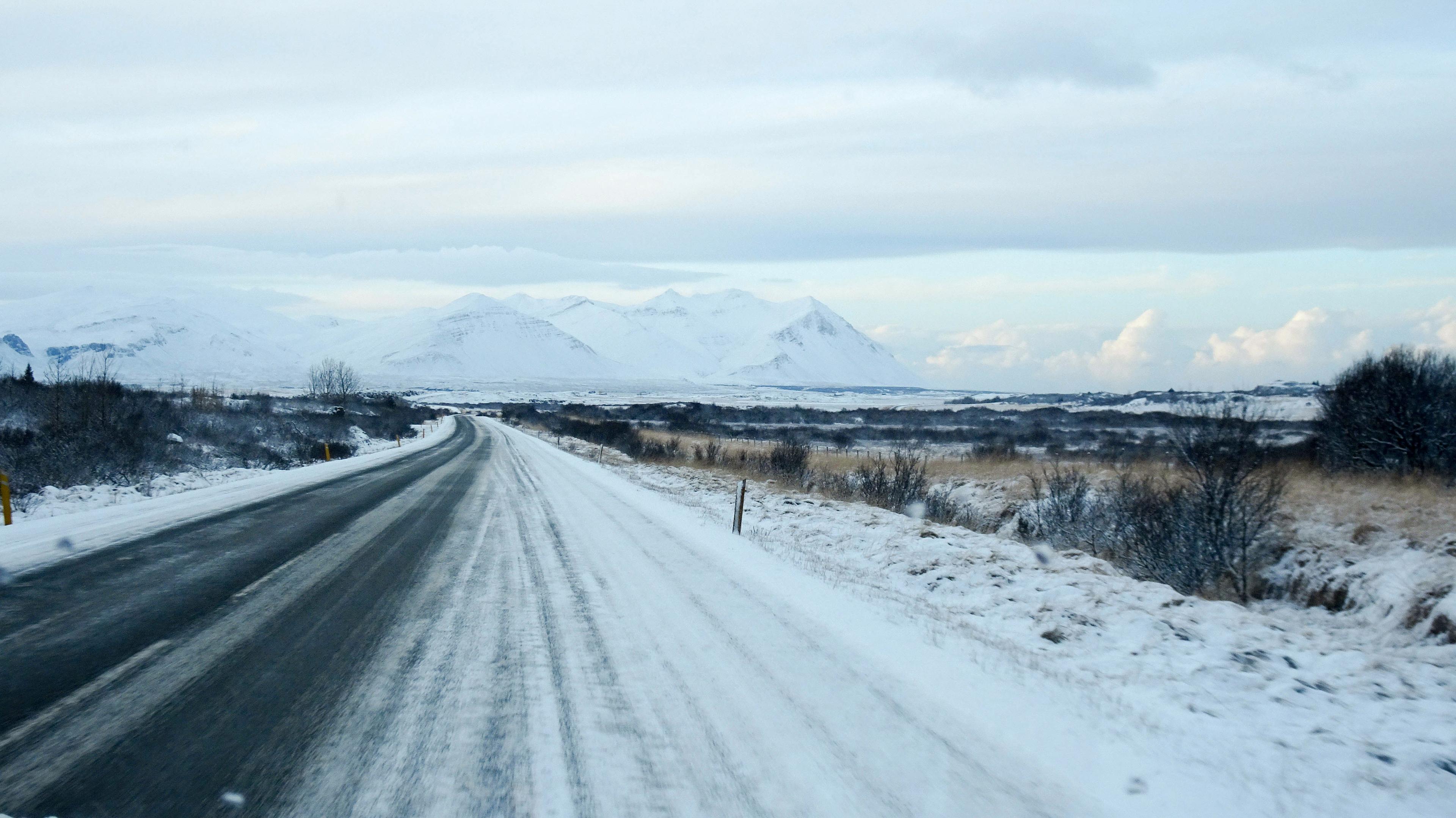 Snow-covered road stretching towards distant snow-capped mountains under a cloudy sky, with bare trees and fields on either side.