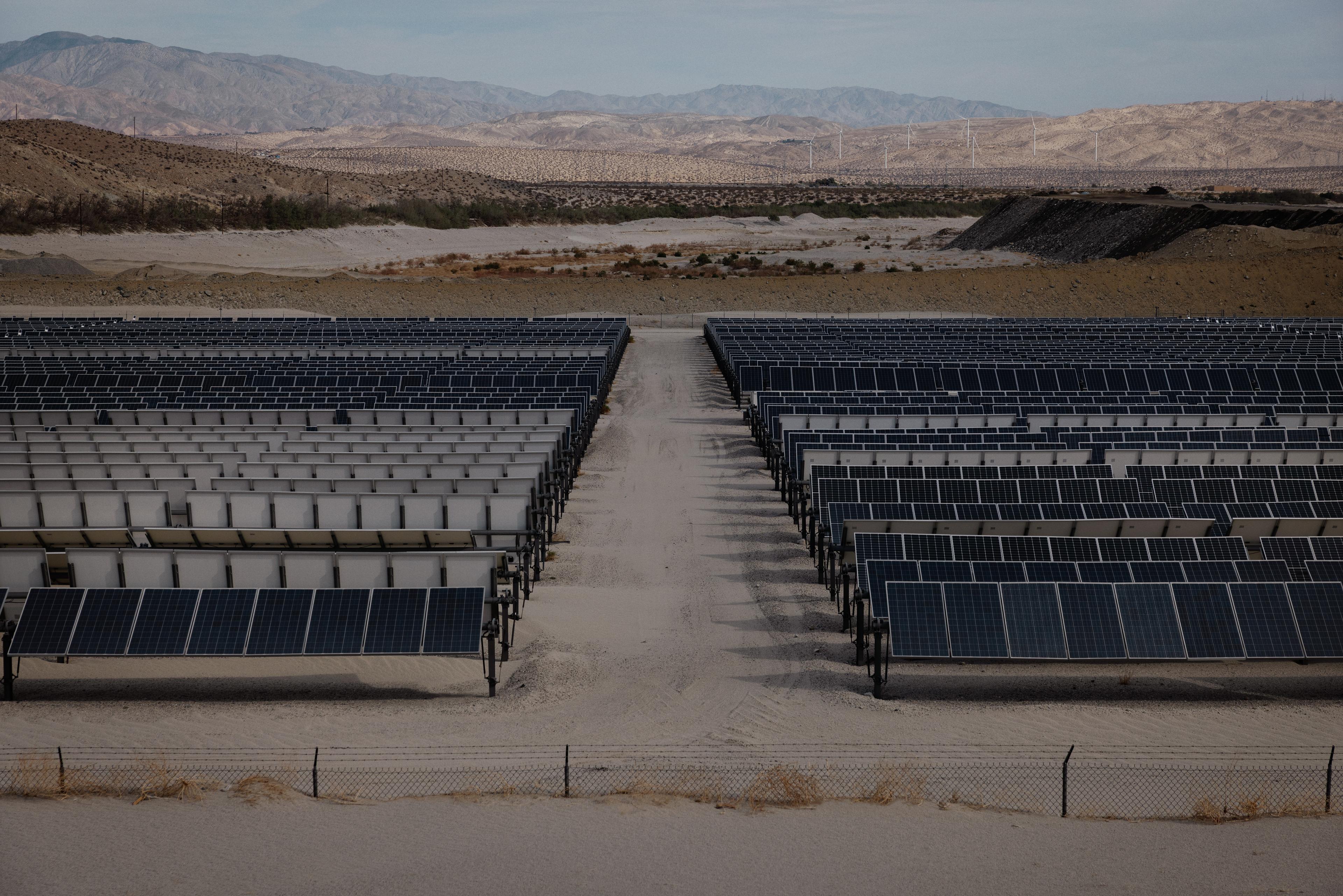 Rows of solar panels in a desert landscape, bordered by mountains under a cloudy sky.