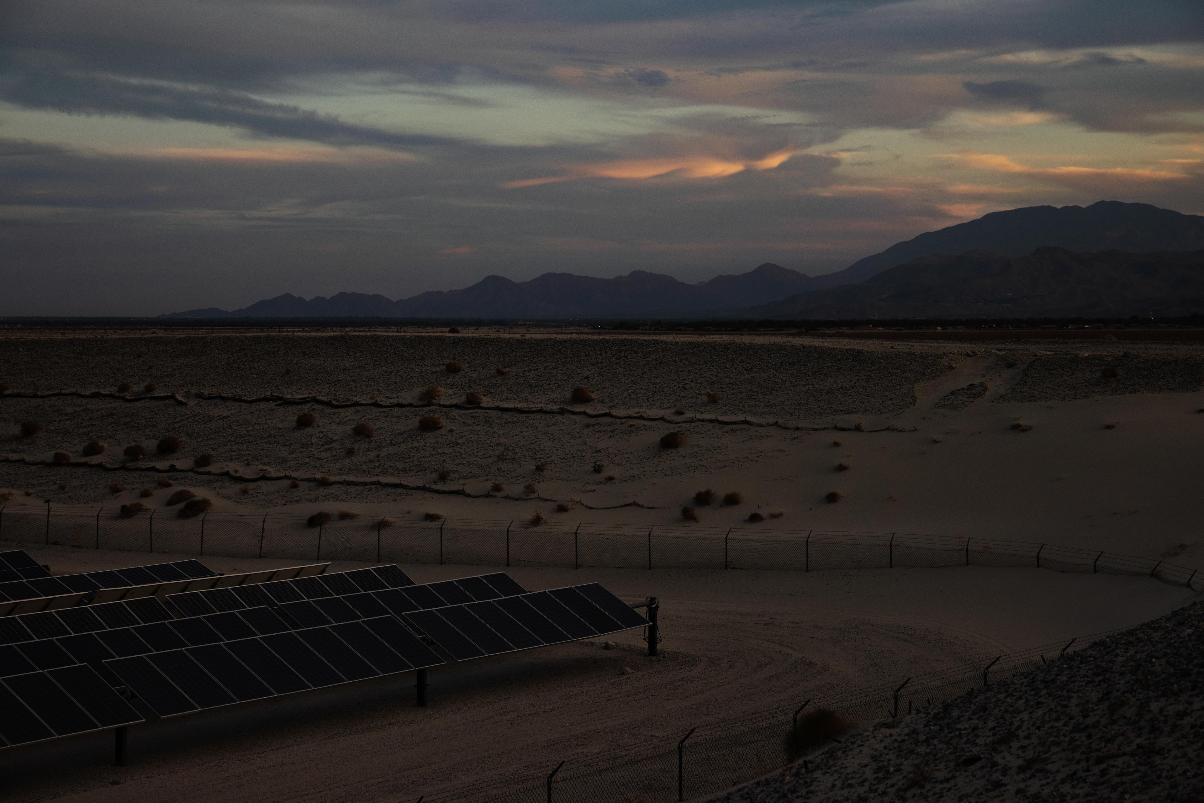 Solar panels in a desert landscape under a cloudy sunset sky, with mountains silhouetted in the background.