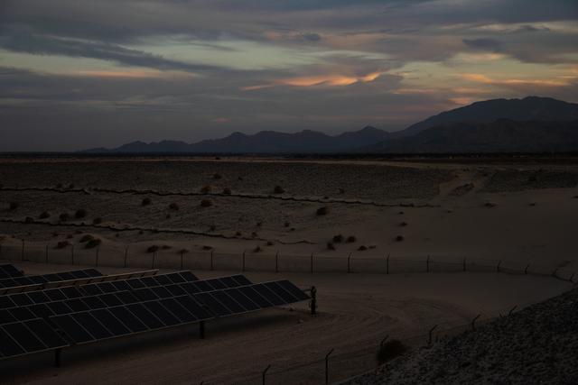 Solar panels in a desert landscape under a cloudy sunset sky, with mountains silhouetted in the background.