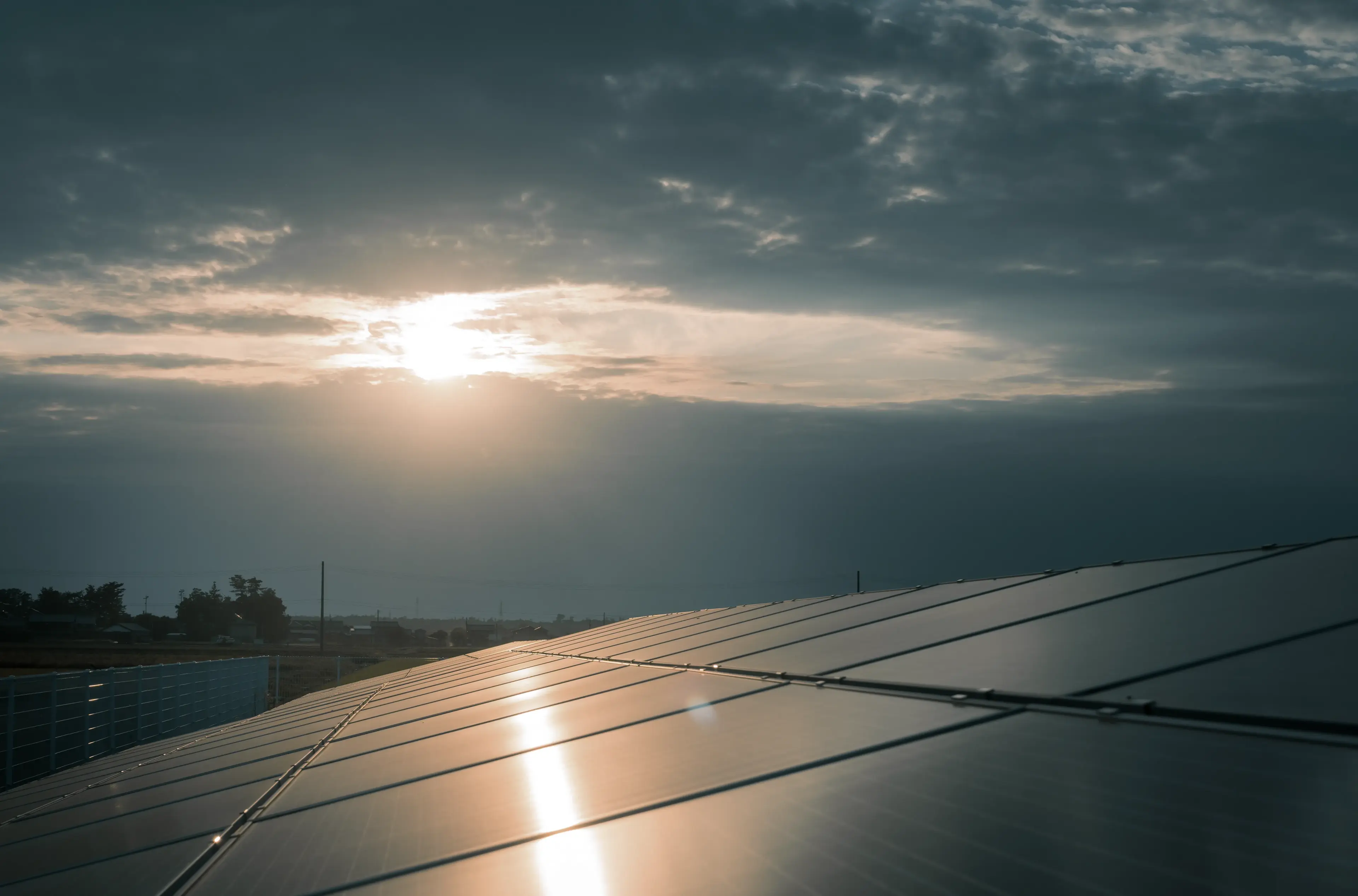 Solar panels under a dark cloudy sky with the sun partially visible, creating a reflective glow on the panels.