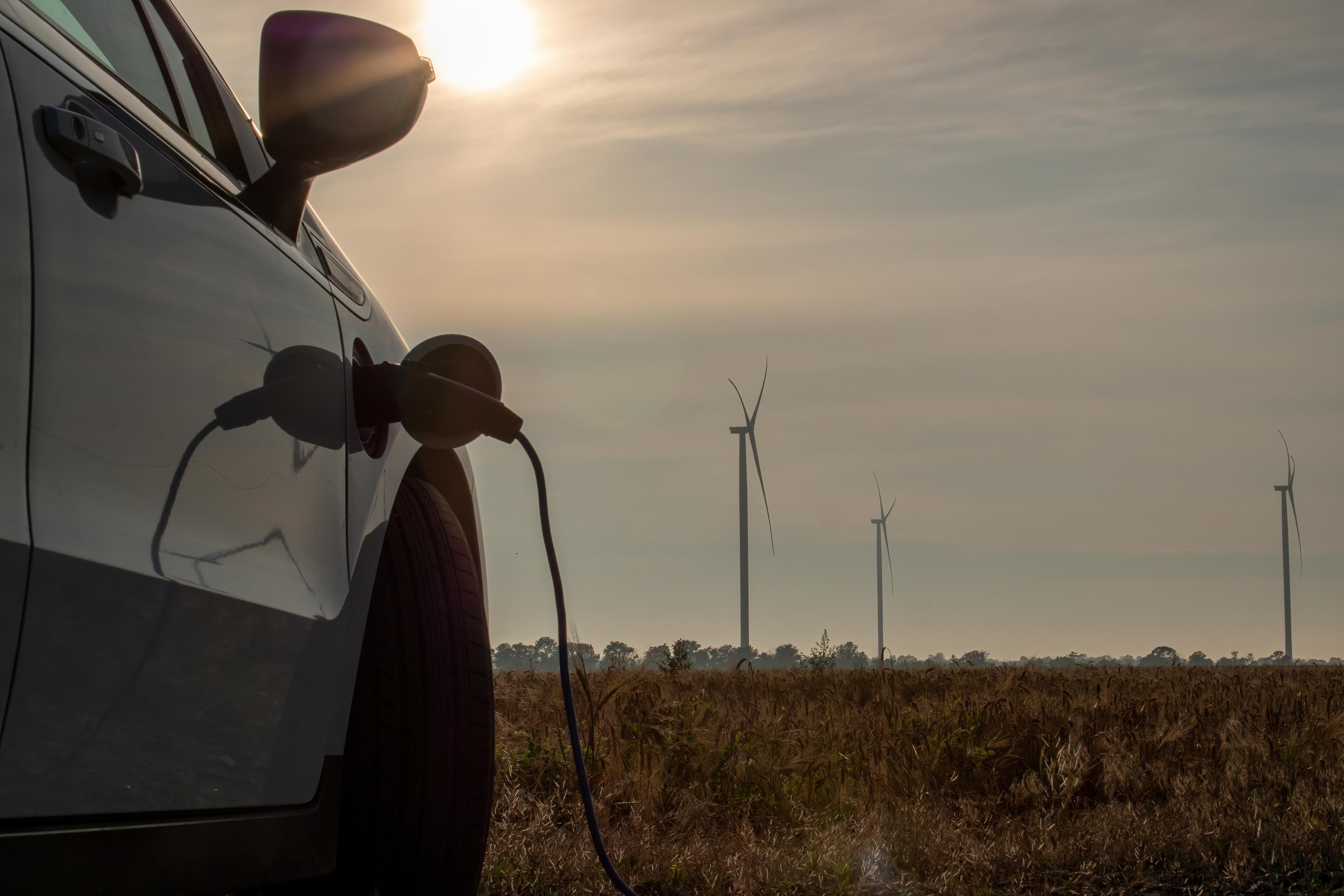 Electric car charging in a rural field with wind turbines in the background under a hazy sky.