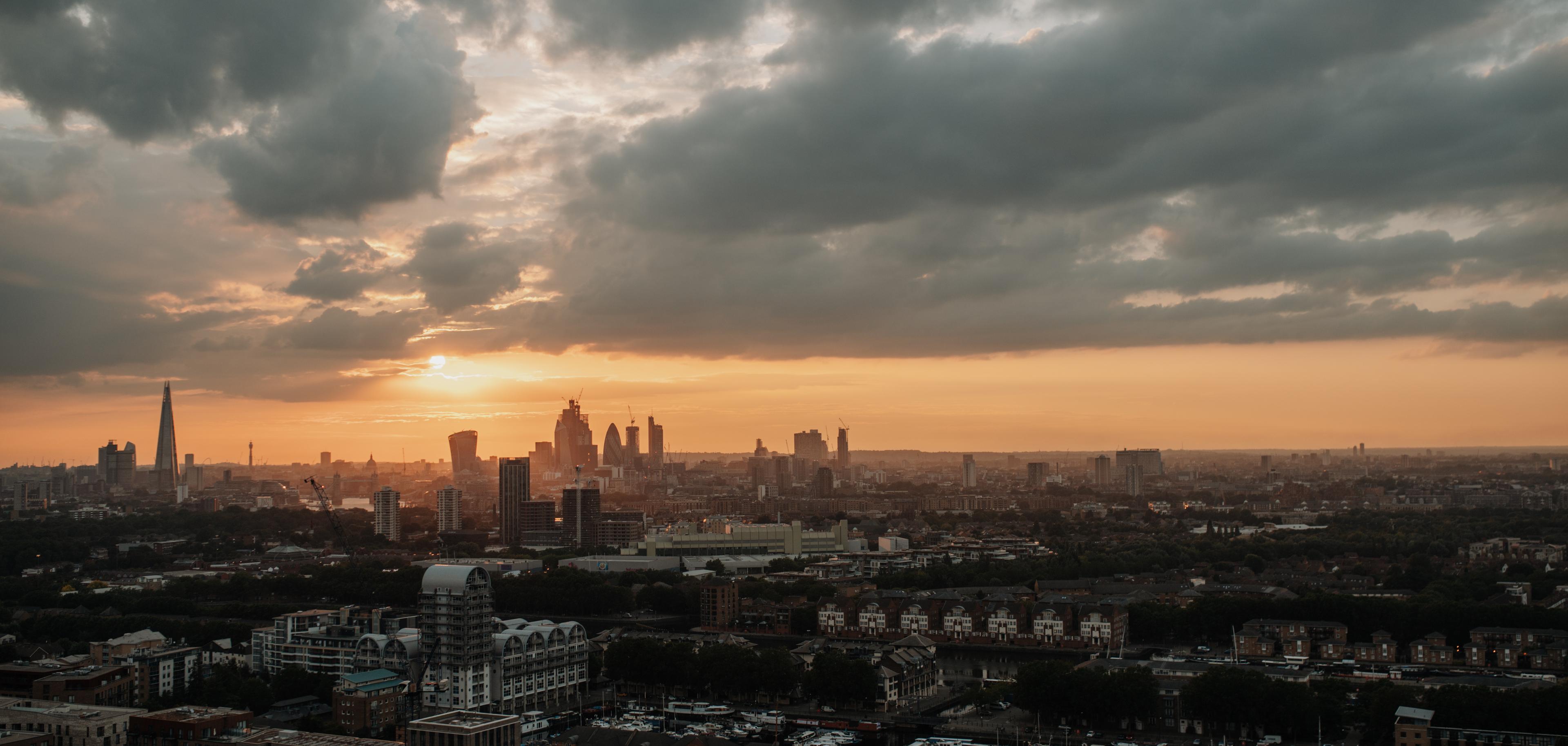 Sunset over a city skyline with tall buildings silhouetted, cloudy sky, and an orange glow on the horizon.