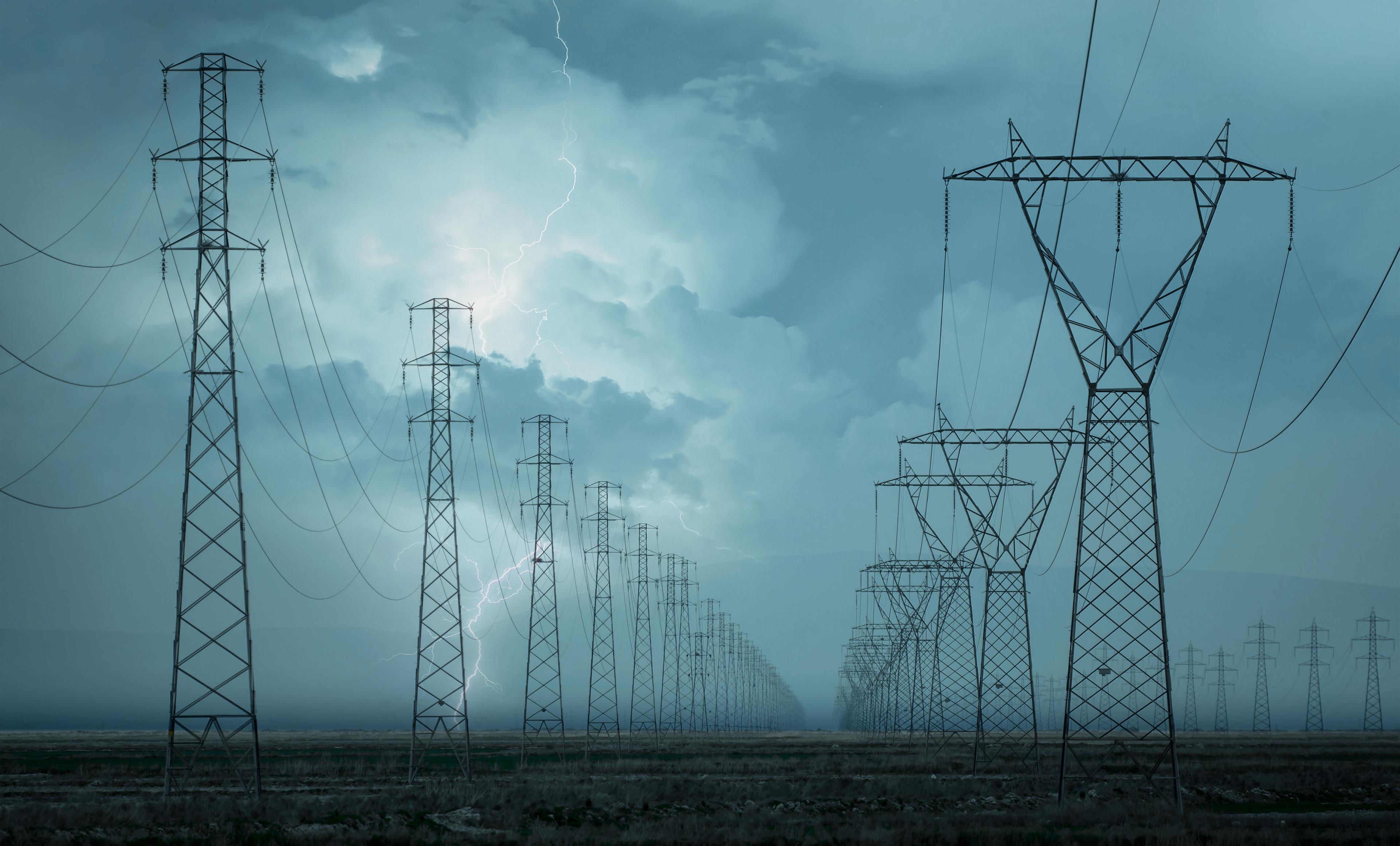 A row of towering power lines in an open field under a stormy sky, with visible lightning in the background.
