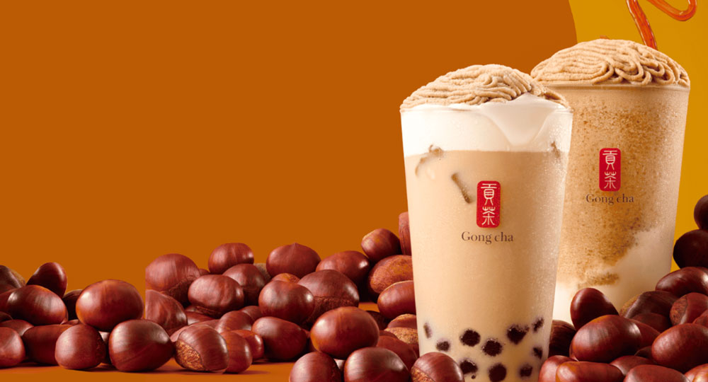 Two bubble tea cups topped with chestnut cream, surrounded by whole chestnuts, against an orange background.