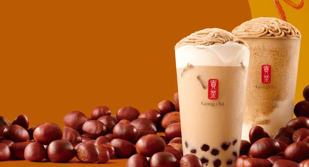 Two bubble tea cups topped with chestnut cream, surrounded by whole chestnuts, against an orange background.