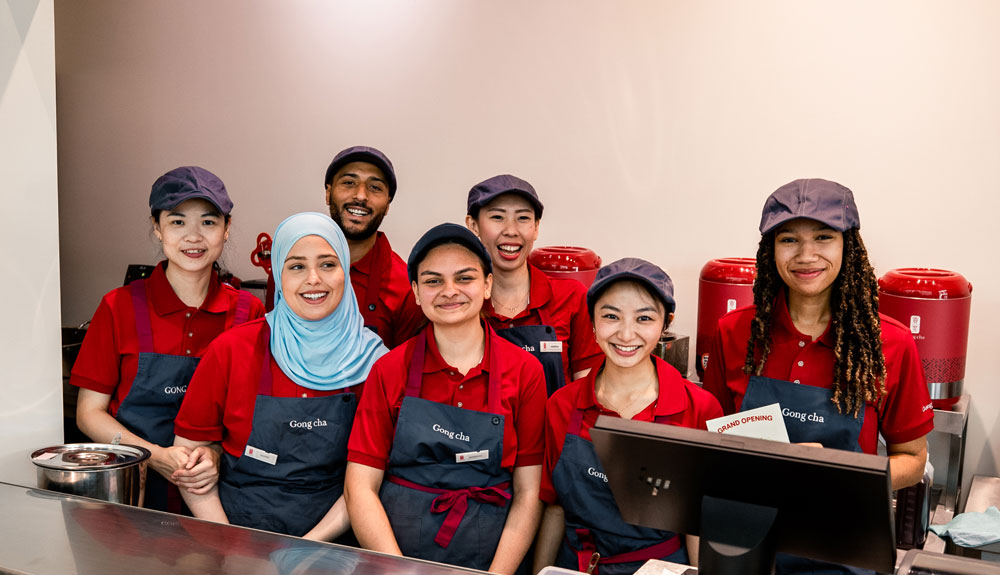 A diverse group of seven smiling employees in red uniforms and navy aprons stand behind a counter, with red dispensers in the background.