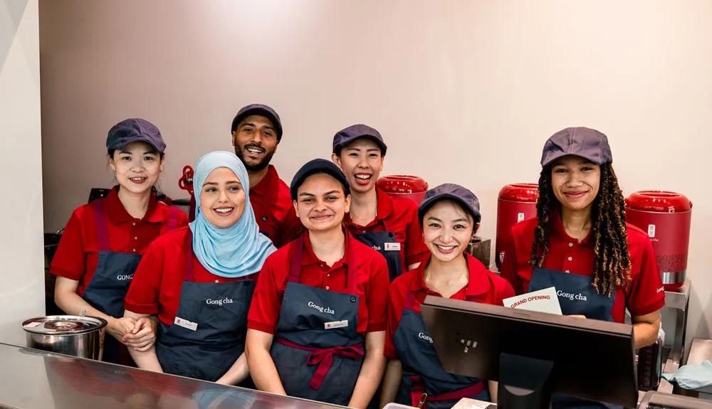 A diverse group of seven smiling employees in red uniforms and navy aprons stand behind a counter, with red dispensers in the background.