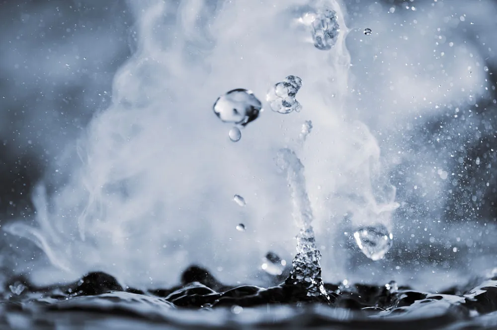 Close-up of water droplets splashing and creating misty steam above a dark liquid surface, with a dynamic and energetic motion captured.