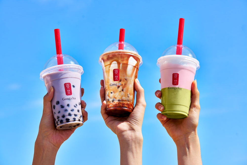 Three hands holding bubble tea cups with red straws against a clear blue sky; flavors include taro, caramel, and strawberry.