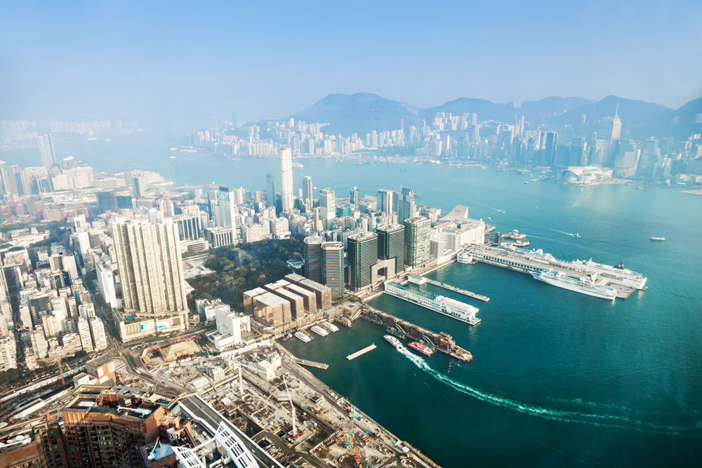 Aerial view of a bustling harbor city with skyscrapers, cruise ships, and mountains in the background under a clear blue sky.
