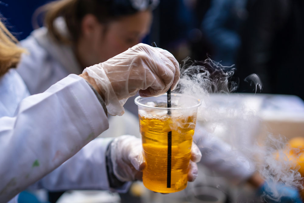 Person in gloves stirring a smoking, amber-colored drink with a black straw, likely involving liquid nitrogen, in a plastic cup.
