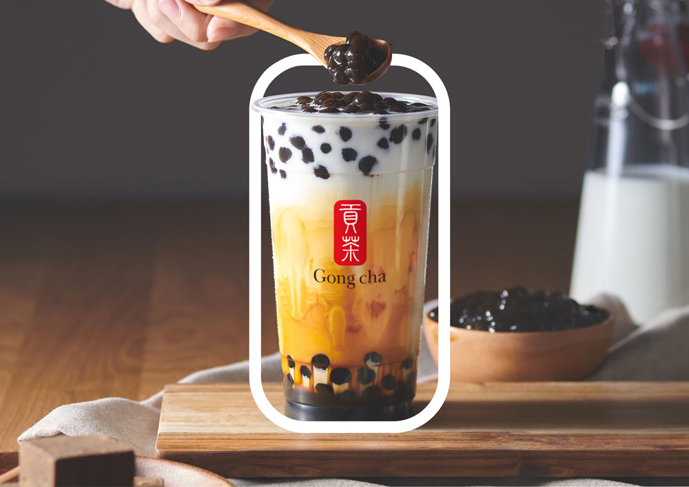 A cup of bubble tea with layers of milk, tea, and tapioca pearls, topped with extra pearls on a wooden tray.