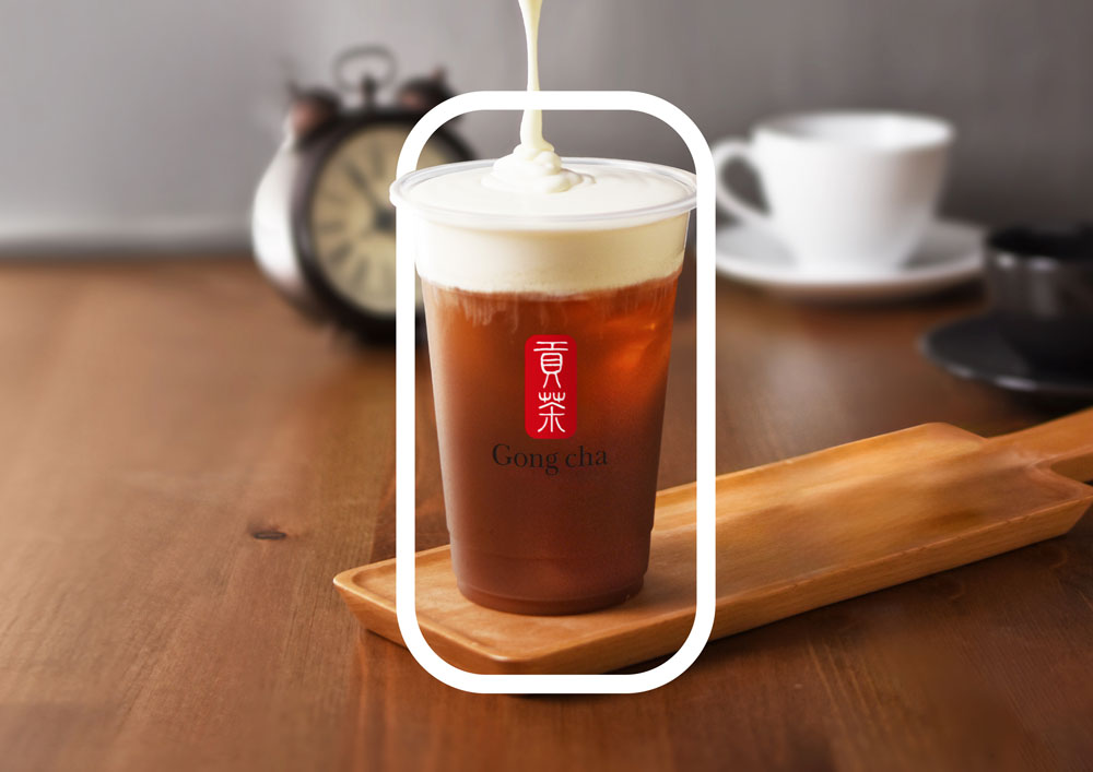 A cup of iced tea with creamy foam on top, placed on a wooden tray. An old-fashioned clock and a white cup are blurred in the background.
