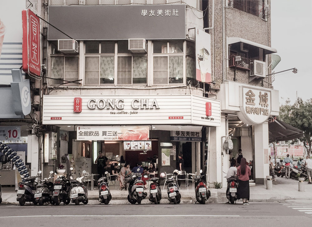 Street view of a corner Gong Cha shop with scooters parked in front and people standing nearby, in an urban setting.