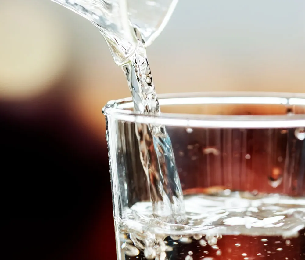 Close-up of water being poured into a clear glass, with bubbles forming on the surface. The background is softly blurred.