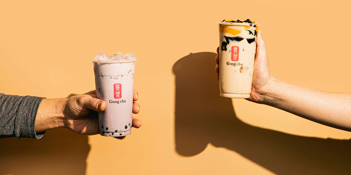 Two hands hold Gong Cha cups — pink milk tea with tapioca pearls and a brown sugar milk tea — against a warm peach backdrop.