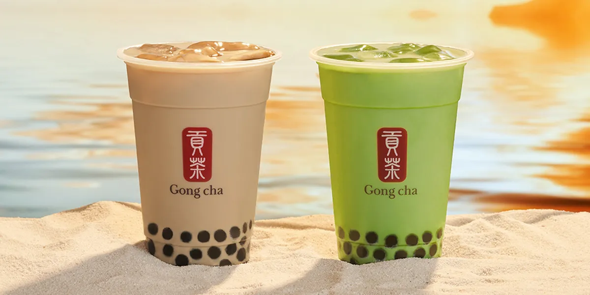Two Gong Cha bubble tea cups with black tapioca pearls, milk tea (left) and matcha (right), on a sandy shore at sunset.