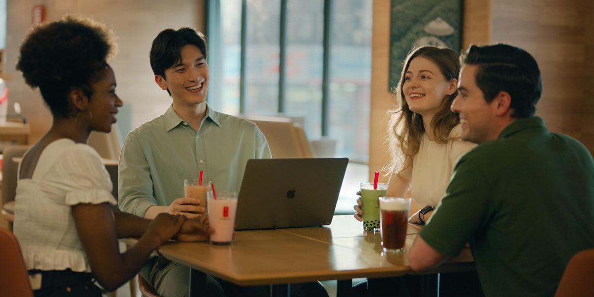Four friends at a café table with laptops, enjoying colourful bubble teas and lively conversation.