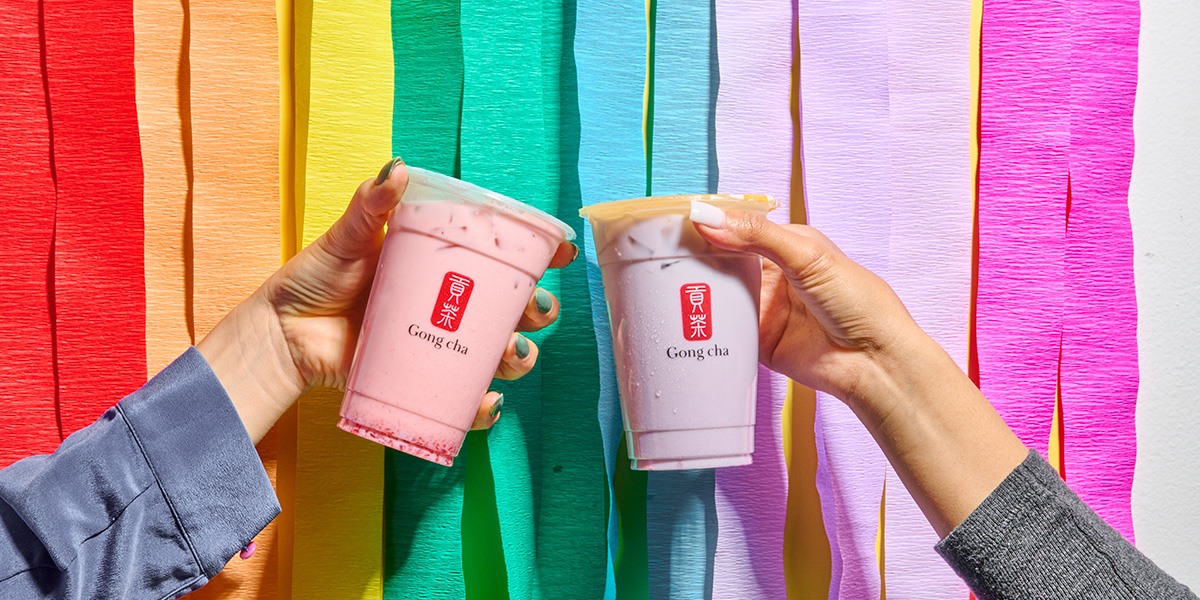 Two hands holding pink Gong Cha bubble tea cups against a rainbow crepe-paper backdrop
