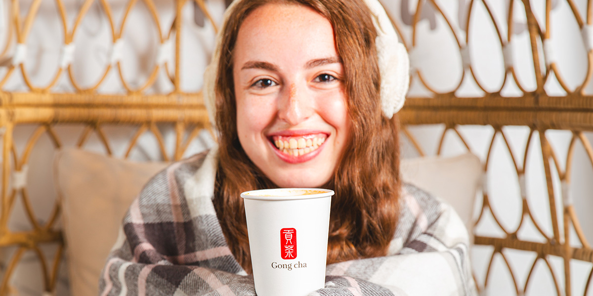 Smiling person wrapped in a plaid blanket holding a Gong Cha cup, seated in a cozy wicker chair.