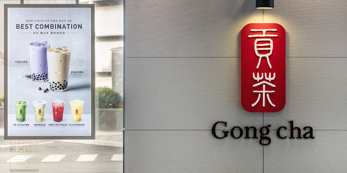 Gong Cha storefront with illuminated red logo plaque and window poster showcasing signature premium bubble tea drinks.