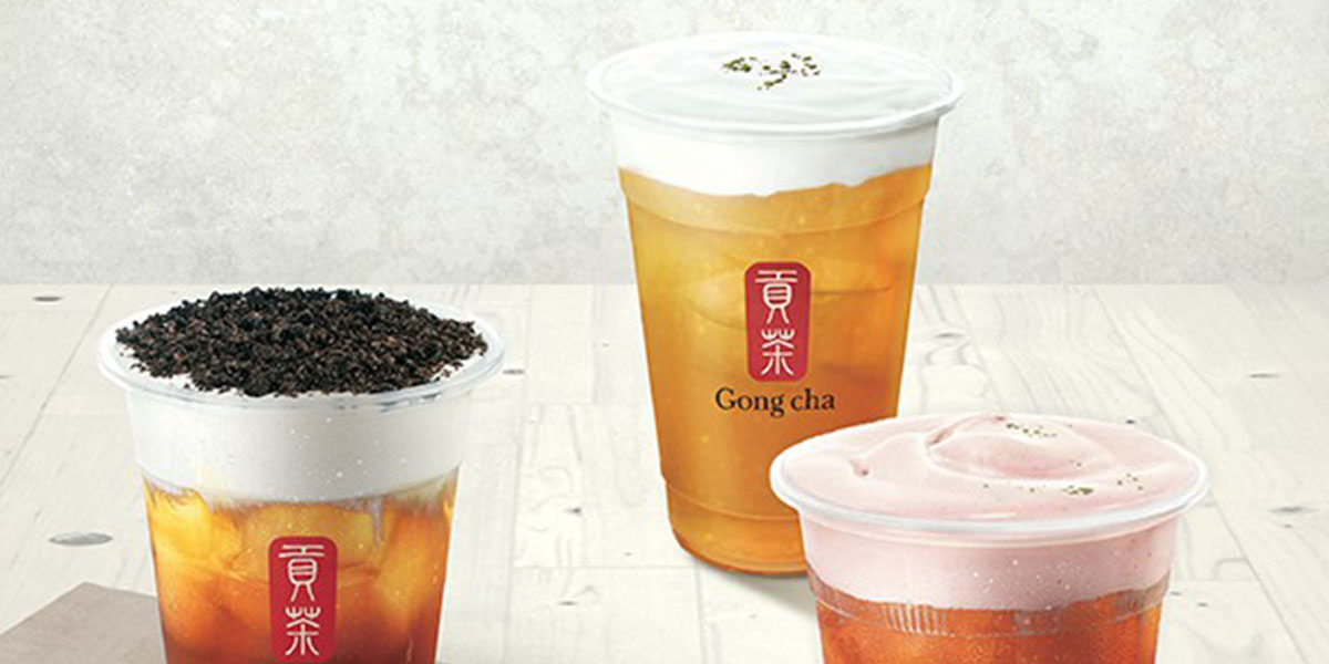 Three Gong Cha iced tea drinks on a light wood table, creamy foam tops with varied toppings, Gong Cha logo visible on cups.