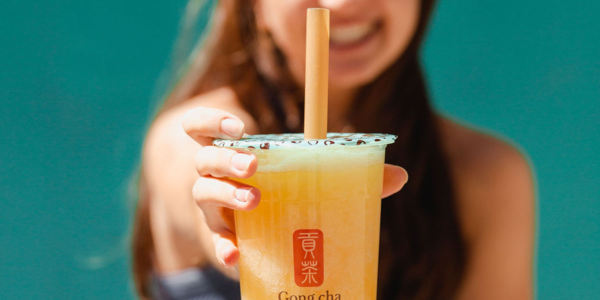 Hand holding a Gong Cha cup of bright yellow fruit tea with a large straw, smiling person blurred against a teal background.