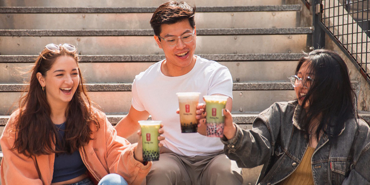 Three friends sitting on stairs laughing and toasting Gong Cha bubble tea cups, two green fruit teas and one brown sugar milk tea.