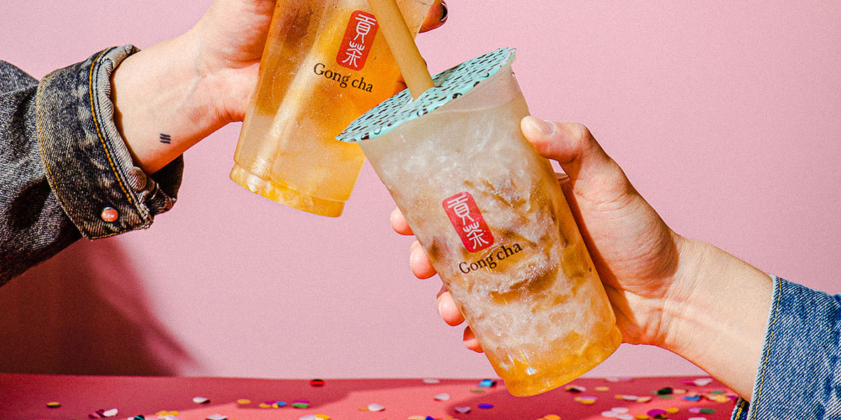 Two hands clinking iced Gong Cha cups with sealed lids over a confetti-speckled pink table.