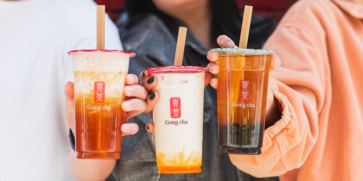 Three hands holding Gong Cha drinks: iced fruit tea, creamy mango milk tea, and tea with boba pearls—customisable toppings visible.