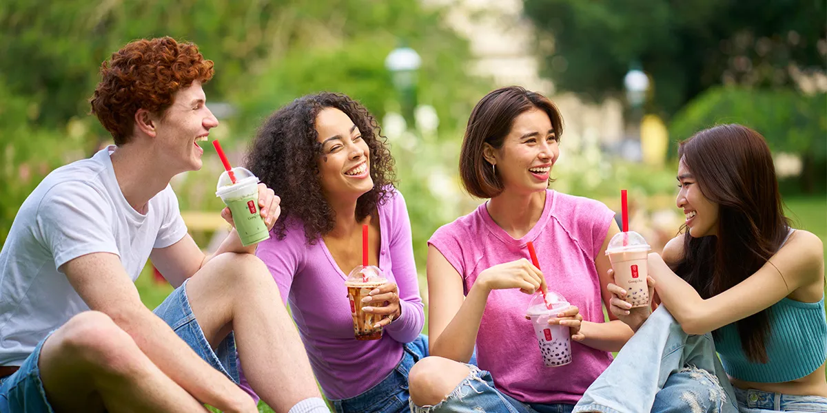 Four friends sit on grass, smiling and enjoying bubble tea with red straws on a sunny day.