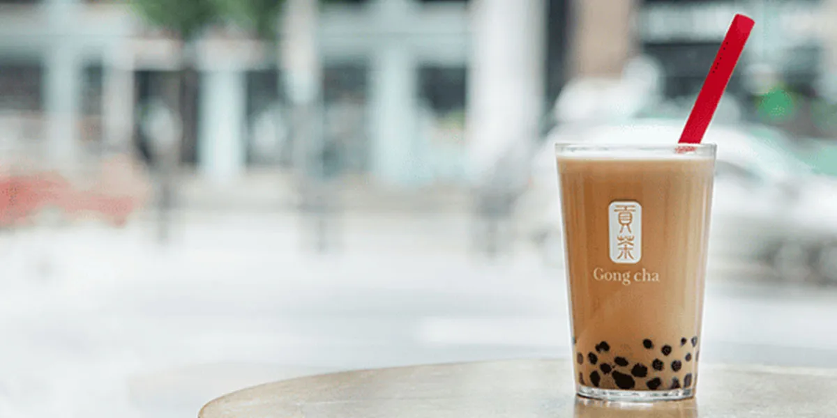 Gong Cha premium milk tea in clear cup with black tapioca pearls and red straw, on a table with blurred urban street background.