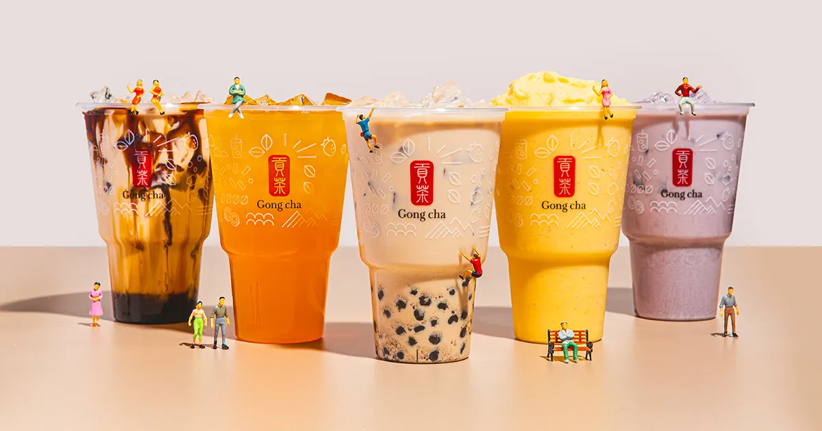 Five Gong Cha bubble tea cups in assorted flavors and toppings, surrounded by tiny toy figurines on a beige surface.