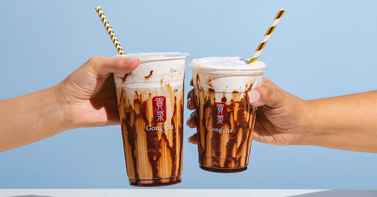 Two hands clinking Gong Cha chocolate-drizzle milk teas with frothy tops and striped paper straws against a pale blue background.